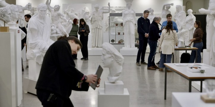 Eine Gruppe an Menschen in einem Saal mit Skulpturen. Eine Frau im Vordergrund erfasst eine Büste mittels eines 3D-Scans über ihr Tablet.