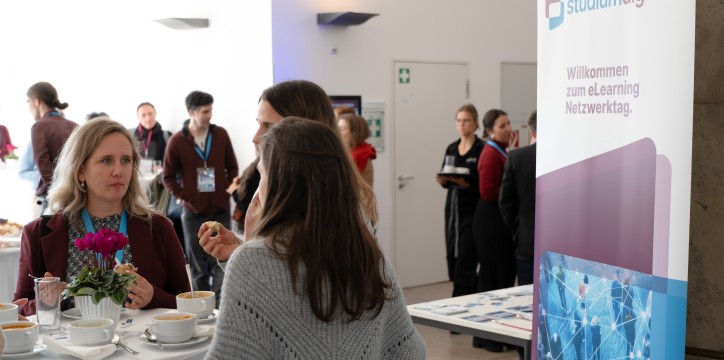 Mehrere Personen stehen in der Mittagspause beisammen bei Essen und Getränken. Auf einem Roll Up im Raum steht der Text „studiumdigitale. Willkommen zum eLearning Netzwerktag“.
