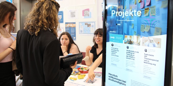 Eine Person mit lockigem Haar, die einen schwarzen Blazer trägt, steht an einem Informationsstand mit dem Titel